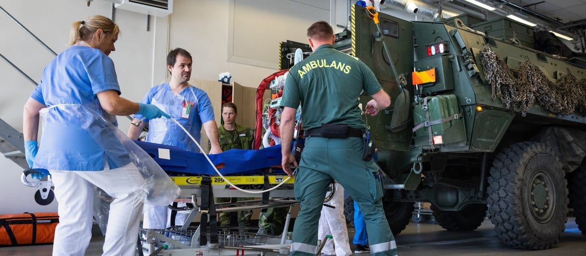 Sjukvårdspersonal, ambulanspersonal och militär personal står vid en bår utanför ett militärt fordon under en samverkansövning.